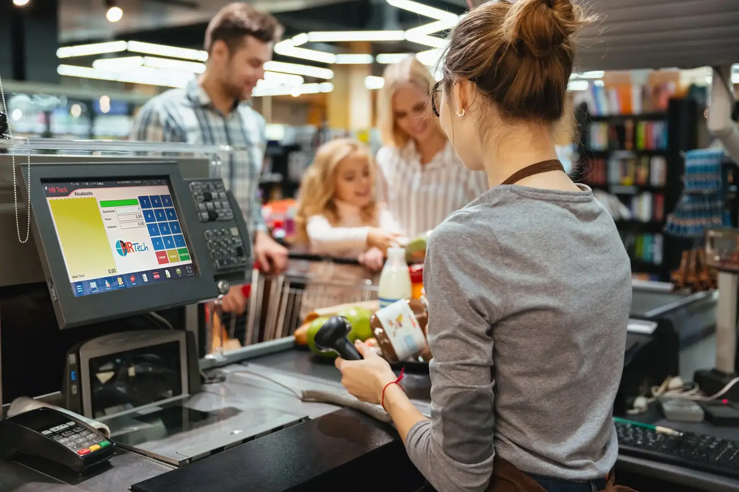 Foto de uma atendente de caixa utilizando o sistema de supermercado da HR Tech para finalizar as compras dos clientes, oferecendo um atendimento ágil, eficiente e agradável aos consumidores.