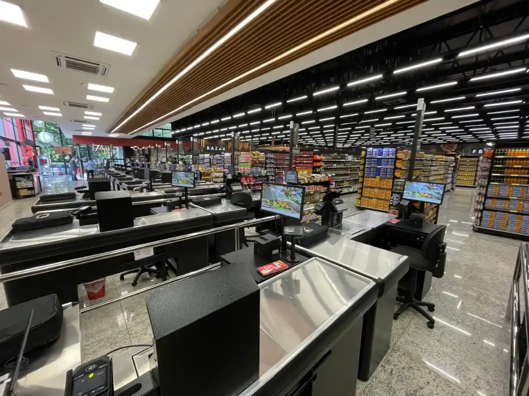 Visão interna de um PDV de supermercado, com caixas registradoras modernas, balcões de atendimento e gôndolas abastecidas ao fundo, em ambiente bem iluminado.
